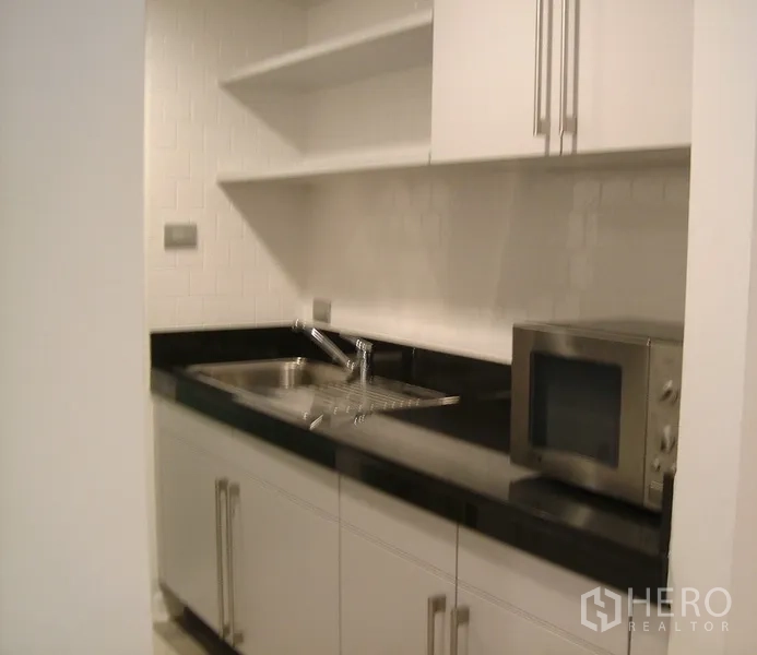 Condominium for rent in Pathum Wan, Bangkok - Compact white kitchen with black stone countertop, sink, open shelves and microwave in a city condominium.