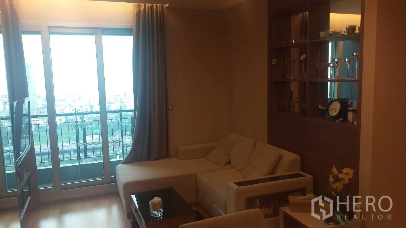 Condominium for rent in Ratchathewi, Bangkok - Cozy living room with L‑shaped sofa, built‑in shelving and balcony doors facing a city skyline.