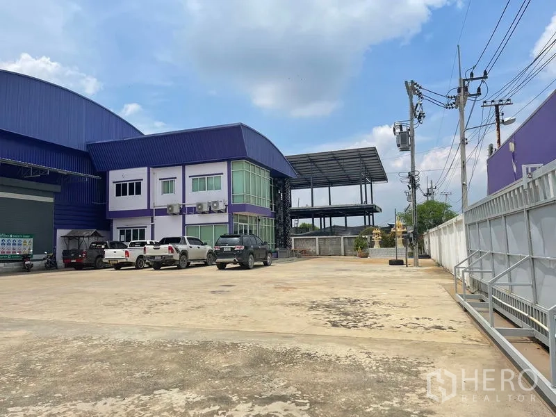 Warehouse for sale in Lam Luk Ka, Pathum Thani - Concrete parking yard beside the two-storey office building with green-tinted windows and a steel-framed covered area to the rear.