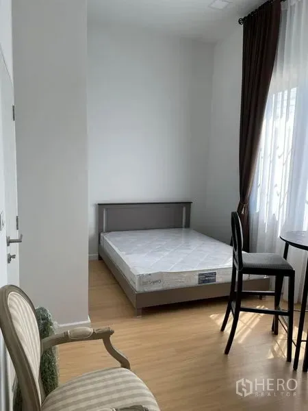 Townhouse for rent in Samut Prakan - Bright bedroom with bed and mattress, tall curtains, small round table and chair on wood flooring.