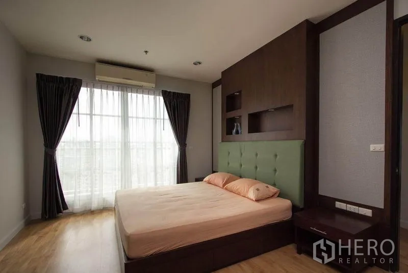 Condominium for rent in Khlong Toei, Bangkok - Master bedroom with padded headboard, wood feature wall, and floor-to-ceiling windows with drapes.