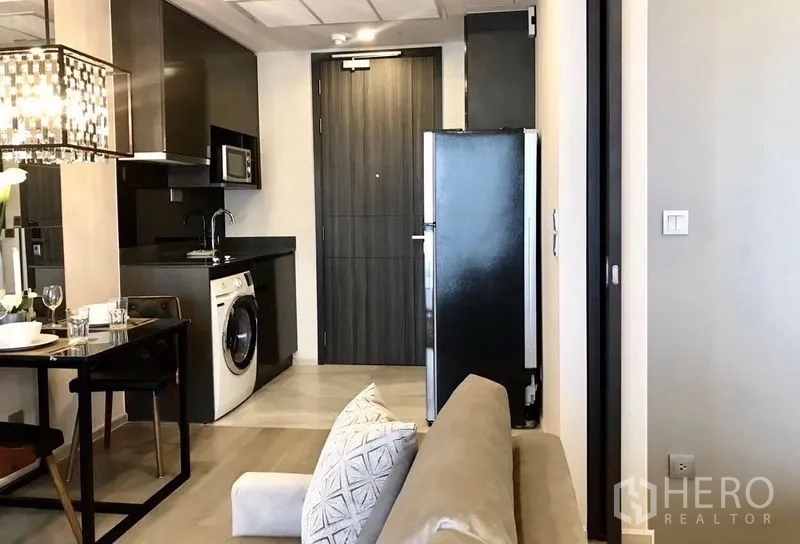 Condominium for rent in Watthana, Bangkok - Compact kitchenette with black cabinets, fridge and in-unit washing machine beside dining table.