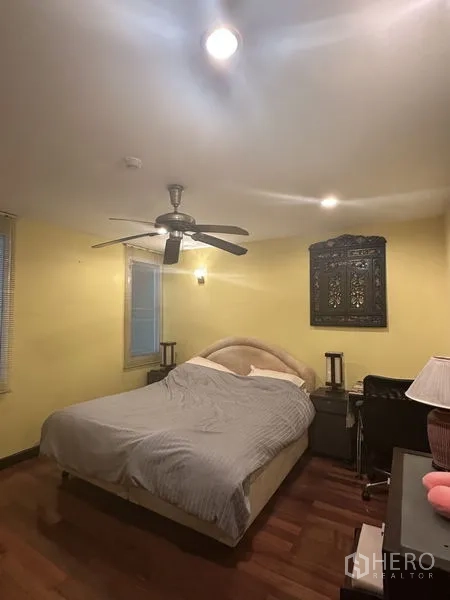 Condominium for sale in Watthana, Bangkok - Cozy bedroom with queen bed, ceiling fan, yellow walls and wood flooring, with bedside tables and desk.