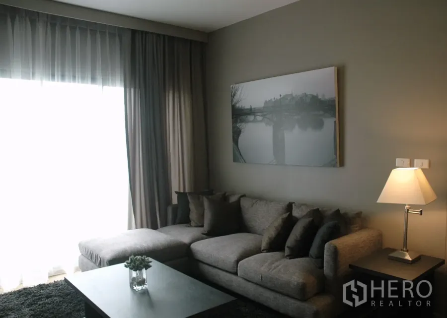 Condominium for rent in Watthana, Bangkok - Cozy living room with gray sectional sofa, floor-to-ceiling curtains, wall art and a table lamp.