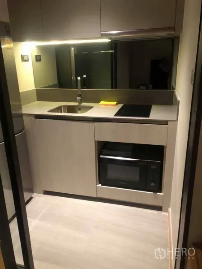 Condominium for rent in Watthana, Bangkok - Compact modern kitchenette with sink, induction cooktop, built-in microwave and mirrored backsplash in an Ekkamai condo.