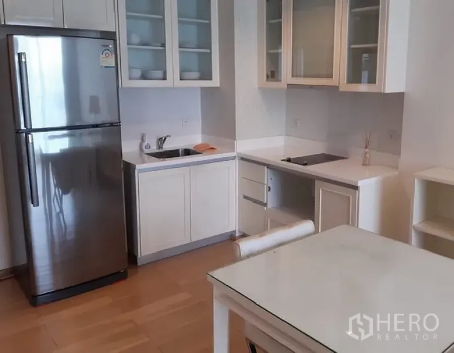 คอนโด ให้เช่าในKhlong Toei, Bangkok - ครัวขนาดกะทัดรัดโทนขาว มีตู้บานกระจก ตู้เย็นสแตนเลส เตาไฟฟ้า และโต๊ะทานอาหารบนพื้นไม้