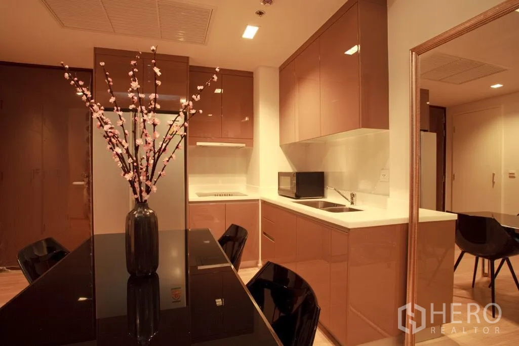 Condominium for rent in Khlong Toei, Bangkok - Modern kitchen with glossy cabinets, solid-surface counters, built-in hob, double sink and dining table.