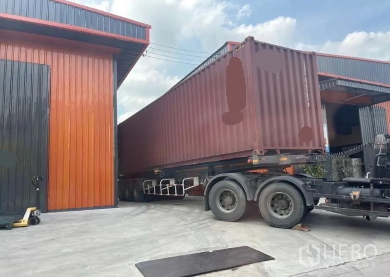 Warehouse for rent in Wang Thonglang, Bangkok - Container trailer maneuvering between orange-and-black metal warehouses with a wide concrete loading yard.