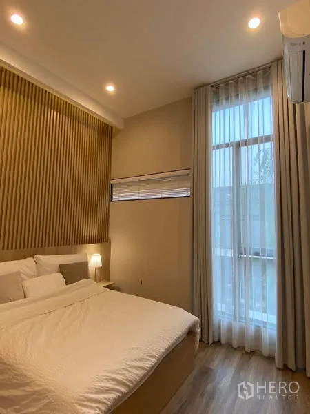 Townhouse for rent in Saphan Sung, Bangkok - Cozy bedroom with wood accent wall, floor-to-ceiling curtains, warm lighting and an air conditioner.