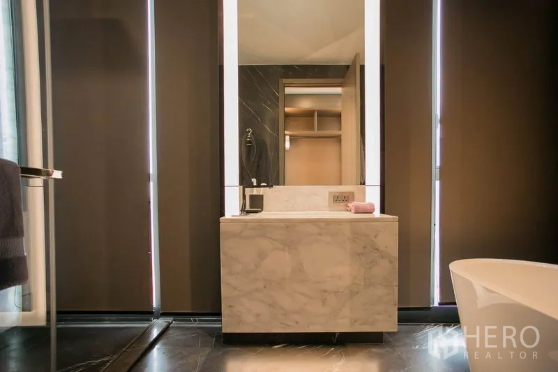 Condominium for rent or sale in Watthana, Bangkok - Bathroom vanity with illuminated mirror, marble sink and blinds beside freestanding tub.