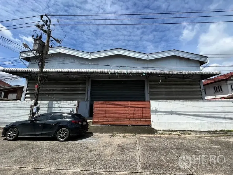 Warehouse for rent in Min Buri, Bangkok - Street view of a gated warehouse with metal awning and large roller shutter in Bangkok.