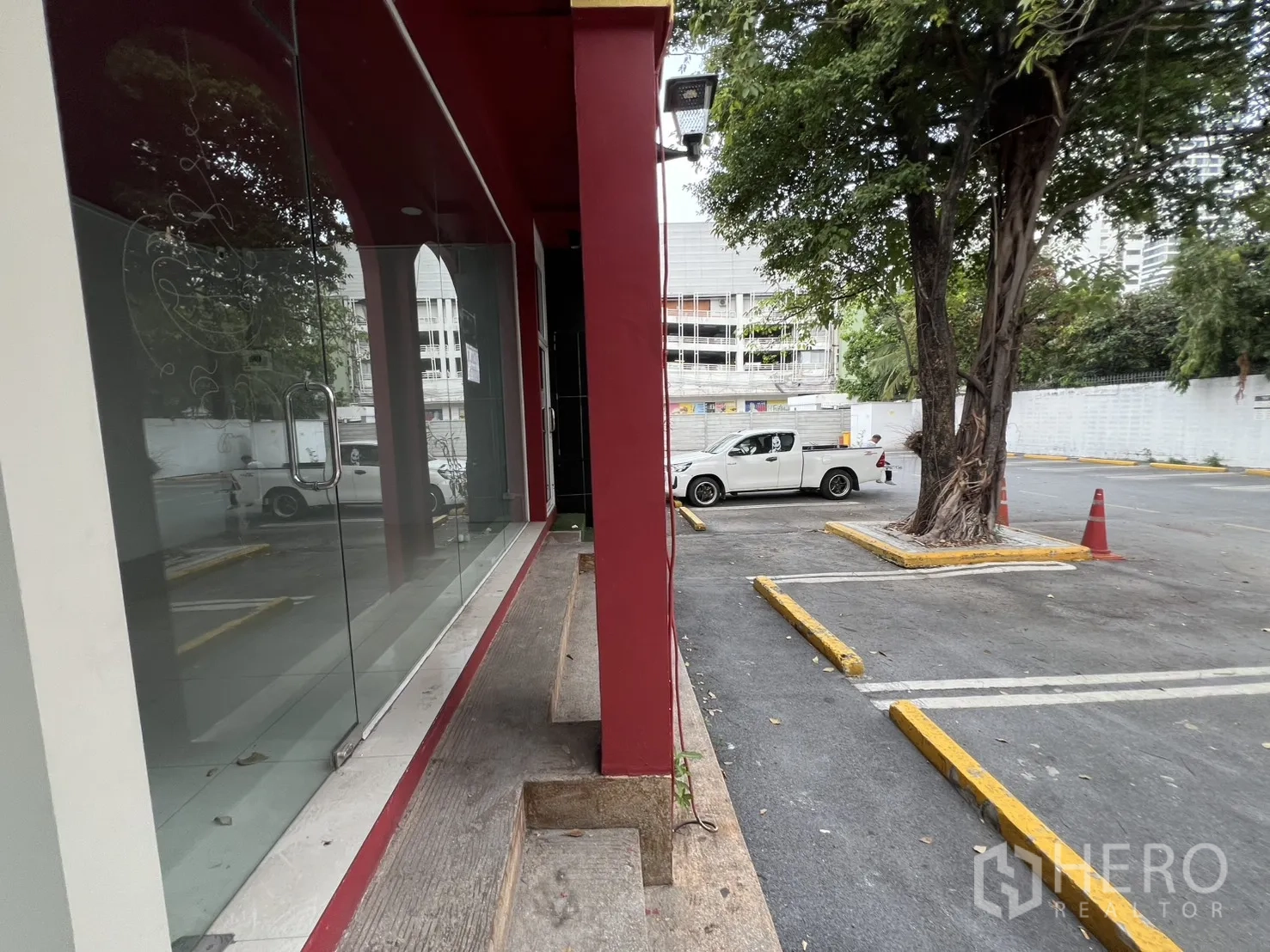 零售空间 for rent in Watthana, Bangkok - 玻璃橱窗旁步道通向开放式停车场，一侧有大树遮阴。