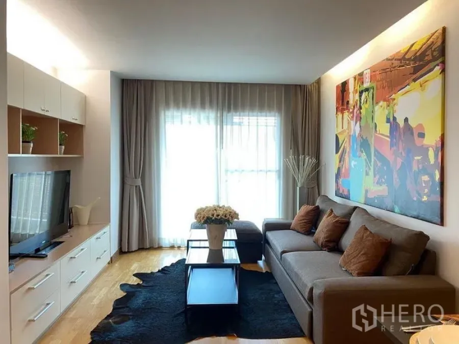 Condominium for sale in Phra Khanong, Bangkok - Living room with sofa, TV cabinet, large artwork, and balcony window with sheer curtains.