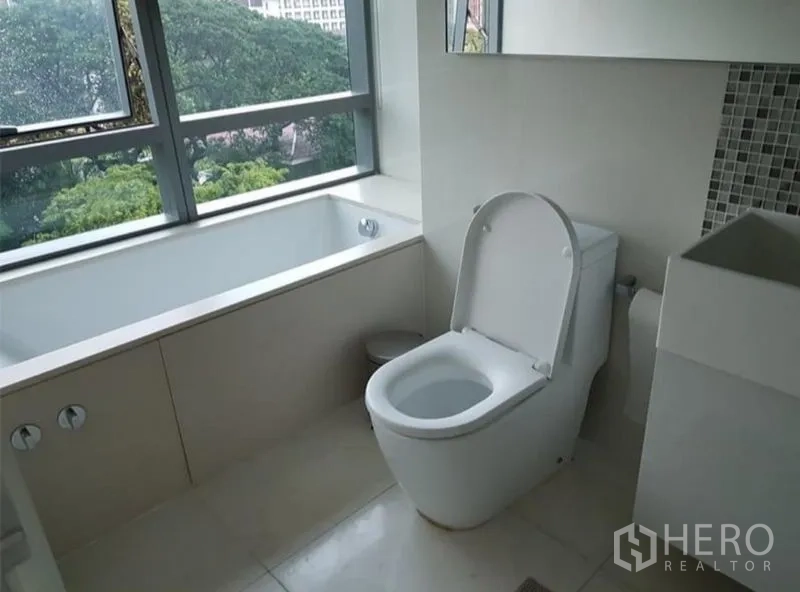 Condominium for rent or sale in Watthana, Bangkok - Modern bathroom with a bathtub by a large window offering green park views and ample natural light.