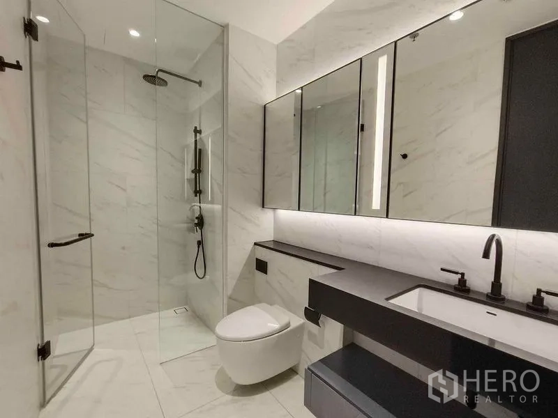 Condominium for rent in Bang Rak, Bangkok - Modern bathroom with glass-enclosed rain shower, wall-mounted toilet, and long vanity.