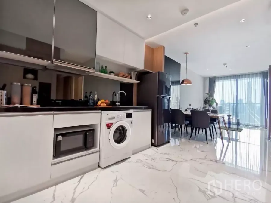 Condominium for rent in Watthana, Bangkok - Modern open‑plan kitchen with washing machine, black countertop and dining area opening to a balcony with city views.