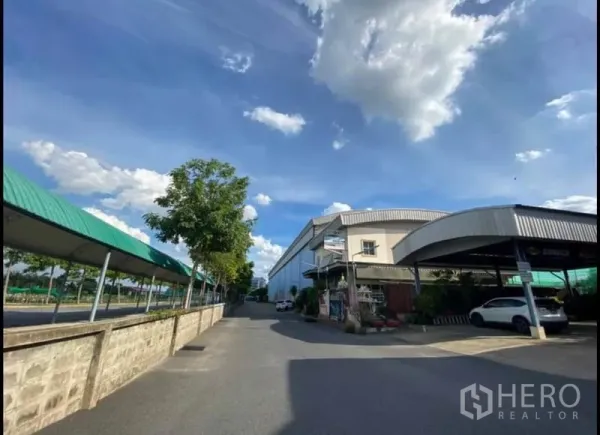 货栈 出租在Sai Mai, Bangkok - 赛迈一条宽阔道路旁的大型蓝色金属外墙仓库，配有遮棚停车位与行道树，天空晴朗。