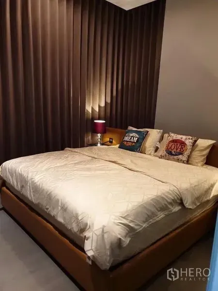 Condominium for rent in Huai Khwang, Bangkok - Bedroom with king-size bed, bedside lamp, and full-height blackout curtains.