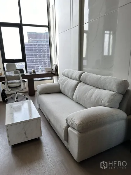 公寓 出租在Bang Rak, Bangkok - 采光充足的角落，灰色沙发与临窗书桌，眺望天际线。