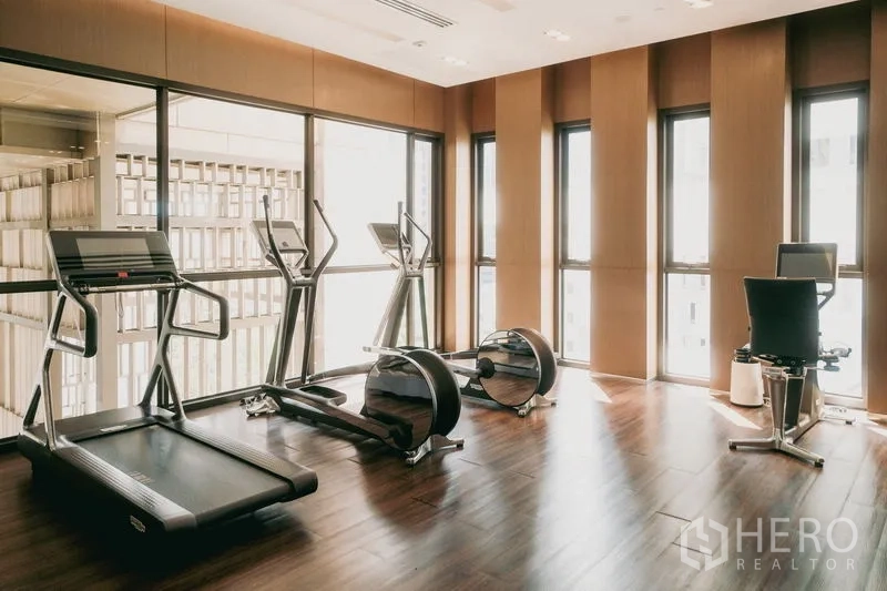 Condominium for rent or sale in Watthana, Bangkok - Fitness room with treadmills and cardio machines on wood floors beside tall windows.