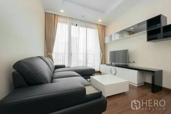 Condominium for rent in Sathon, Bangkok - Living room with black leather sectional sofa, TV console and large curtained window.
