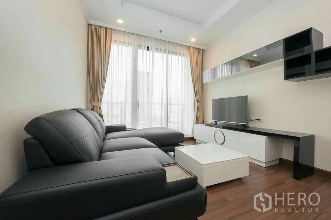 Condominium for rent in Sathon, Bangkok - Living room with black leather sectional sofa, TV console and large curtained window.