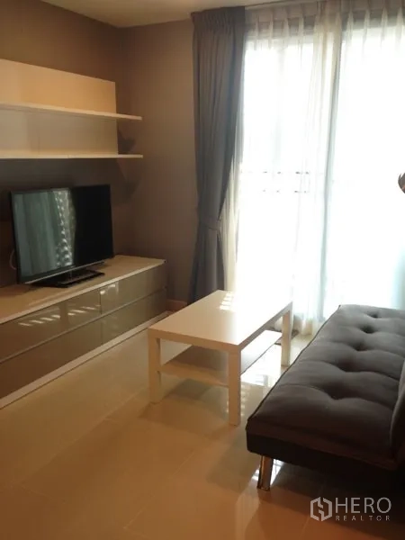 Condominium for rent or sale in Mueang Samut Prakan, Samut Prakan - Cozy living room with glossy TV unit, gray sofa, white coffee table and balcony door.