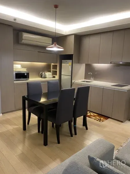 Condominium for rent in Watthana, Bangkok - Open kitchen with built‑in cabinetry, cooktop, fridge, and a dining table for four beneath a pendant light.