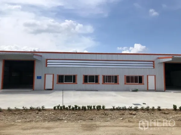 Warehouse for rent in Nonthaburi - Modern metal-clad warehouse facade with orange trim, twin wide roller doors and a large concrete loading yard under a clear sky in Nonthaburi.