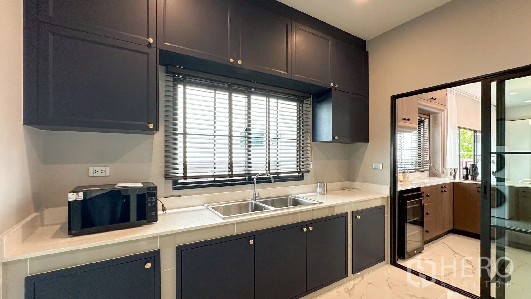 Detached House for rent in Bang Phli, Samut Prakan - Bright kitchen with dark blue cabinets, twin sinks beneath a window, and a sliding door to the pantry.