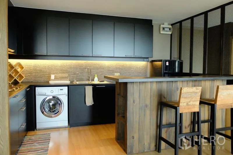 Condominium for sale in Watthana, Bangkok - Kitchen featuring a wooden island with two bar stools and sleek black cabinetry.