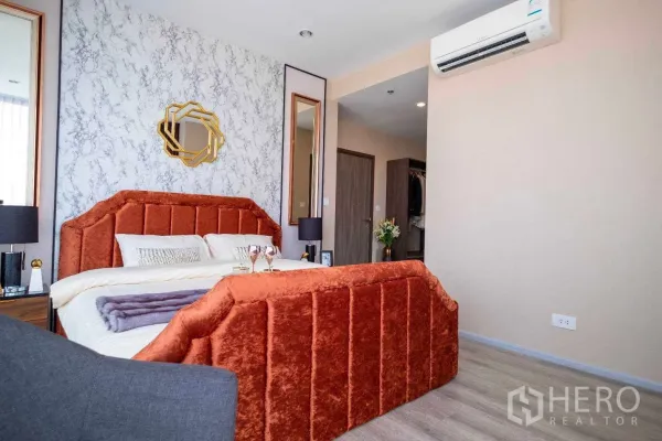 คอนโด ให้เช่าในBang Na, Bangkok - มุมมองห้องนอนมีเตียงบุผ้าสีส้ม ผนังลายหินอ่อน กระจกเงาคู่ และเครื่องปรับอากาศต่อเนื่องไปยังโซนแต่งตัว