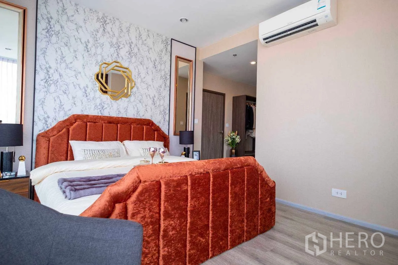 คอนโด ให้เช่าในBang Na, Bangkok - มุมมองห้องนอนมีเตียงบุผ้าสีส้ม ผนังลายหินอ่อน กระจกเงาคู่ และเครื่องปรับอากาศต่อเนื่องไปยังโซนแต่งตัว