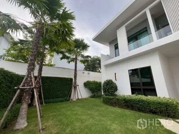 Detached House for rent in Samut Prakan - Lush side garden with palm trees and manicured lawn beside the white facade.