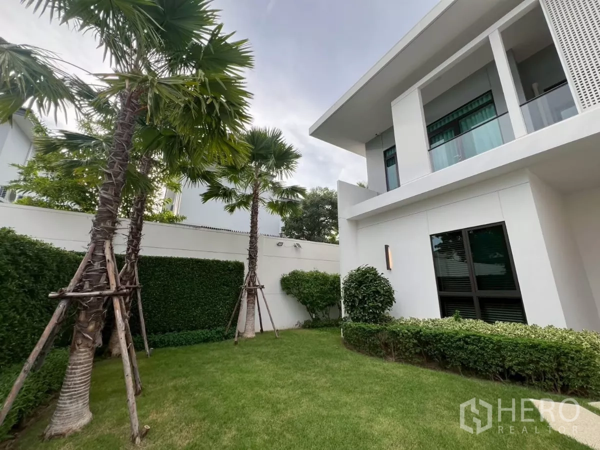 Detached House for rent in Samut Prakan - Lush side garden with palm trees and manicured lawn beside the white facade.