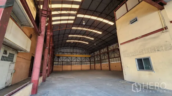 货栈 出租在Nonthaburi - 拱形高屋顶的宽敞仓库内部，带采光带与红色钢柱。