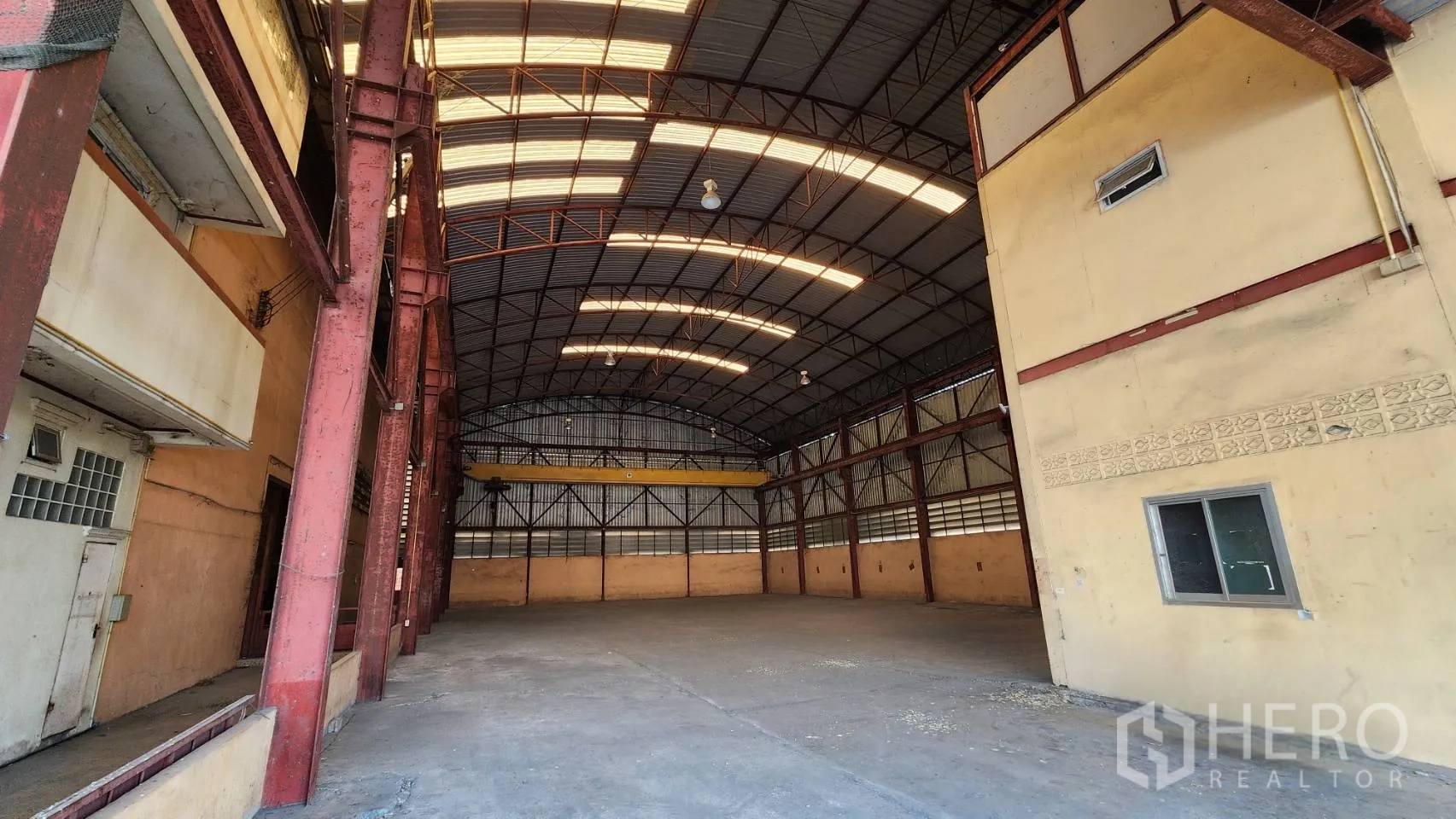 货栈 出租在Nonthaburi - 拱形高屋顶的宽敞仓库内部，带采光带与红色钢柱。