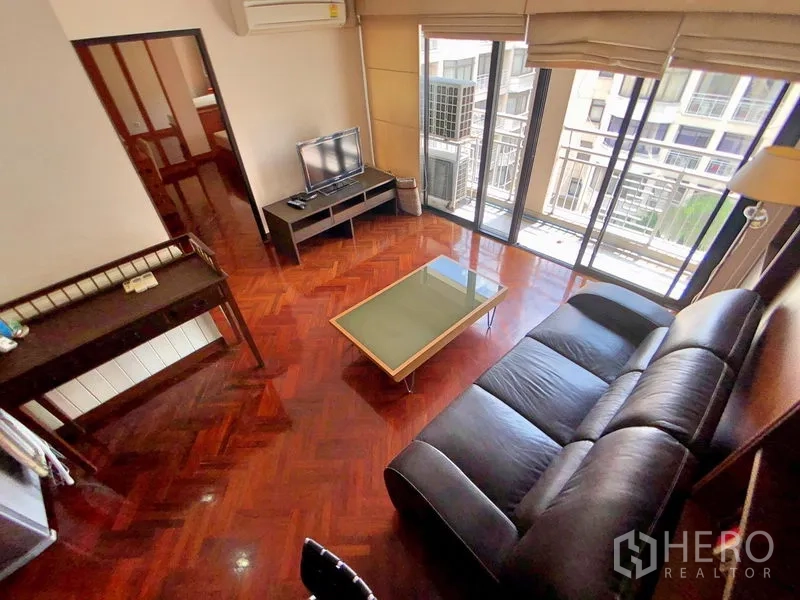 公寓 出租在Bang Rak, Bangkok - 明亮客厅，黑色皮沙发、玻璃茶几与电视柜，落地窗通往阳台，享城市景观。