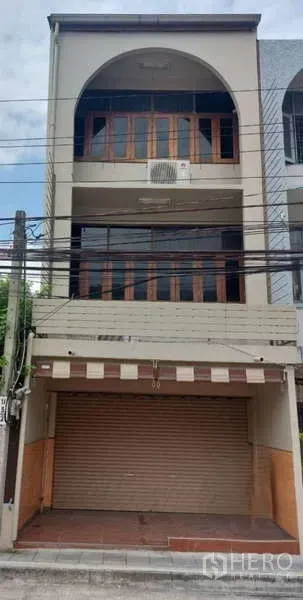 店屋 出租在Phra Khanong, Bangkok - 三层店屋外立面，卷闸门与拱形阳台，上方木框玻璃窗。