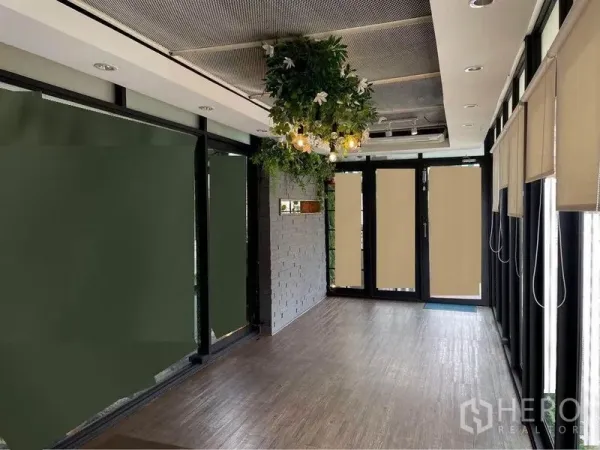 零售空间 出租在Watthana, Bangkok - 明亮的商铺室内，木地板、玻璃门面、悬挂绿植与卷帘。