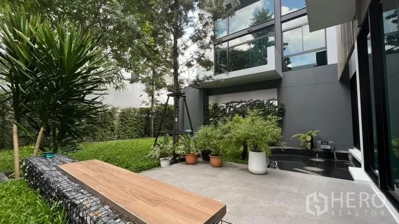 Detached House for sale in Saphan Sung, Bangkok - Landscaped garden patio with bench seating, potted plants and large glass windows of the home.
