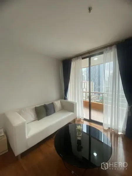 คอนโด ให้เช่าในPathum Wan, Bangkok - ห้องนั่งเล่นสว่างด้วยโซฟาสีขาว โต๊ะกลางทรงกลม และประตูกระจกสไลด์ออกสู่ระเบียงชมวิวสกายไลน์กรุงเทพฯ