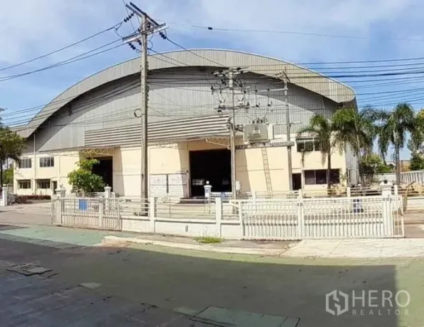 工厂 出租在Mueang Samut Prakan, Samut Prakan - 街景外观：大型弧形屋顶工厂，带大门、棕榈树和架空电线。