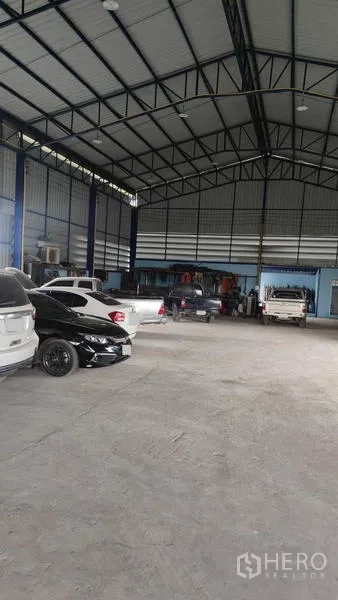 Warehouse for sale in Nakhon Pathom - Spacious warehouse floor with high ceiling, concrete surface and vehicles parked beside a small workshop area.
