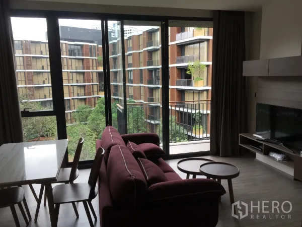 Condominium for rent in Watthana, Bangkok - Living room with maroon sofa and TV facing floor-to-ceiling windows opening to a balcony with lush courtyard views.