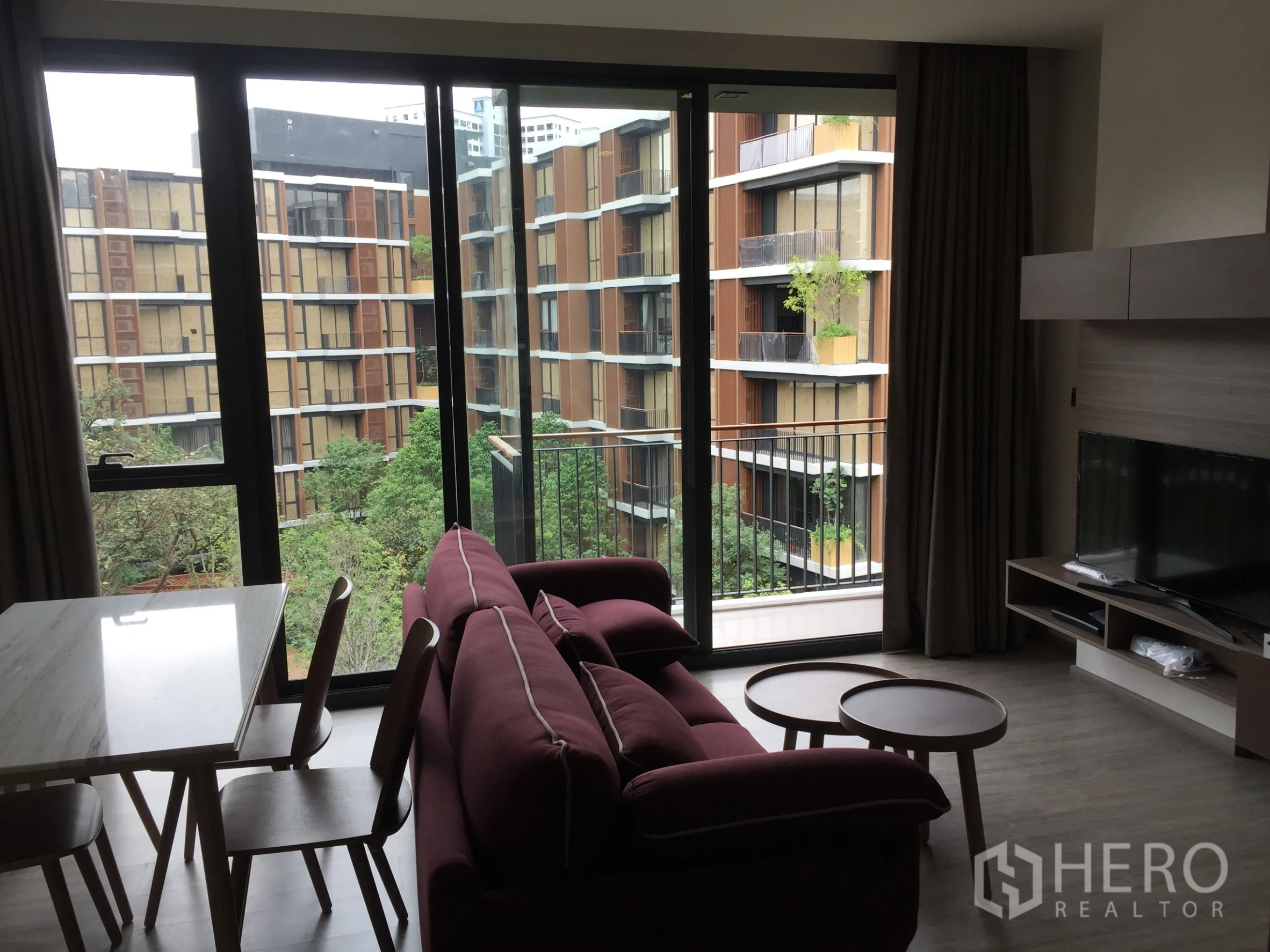 Condominium for rent in Watthana, Bangkok - Living room with maroon sofa and TV facing floor-to-ceiling windows opening to a balcony with lush courtyard views.