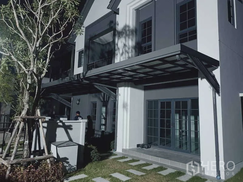 Detached House for rent in Bang Phli, Samut Prakan - Contemporary two-storey house facade at night with large windows, metal awning and small lawn.