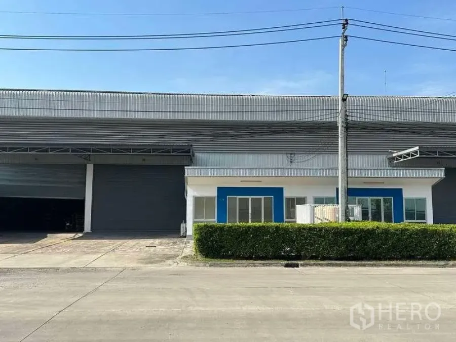 Factory for rent in Bang Sao Thong, Samut Prakan - Modern factory frontage with large roller shutter, blue-accented office, hedge and driveway.