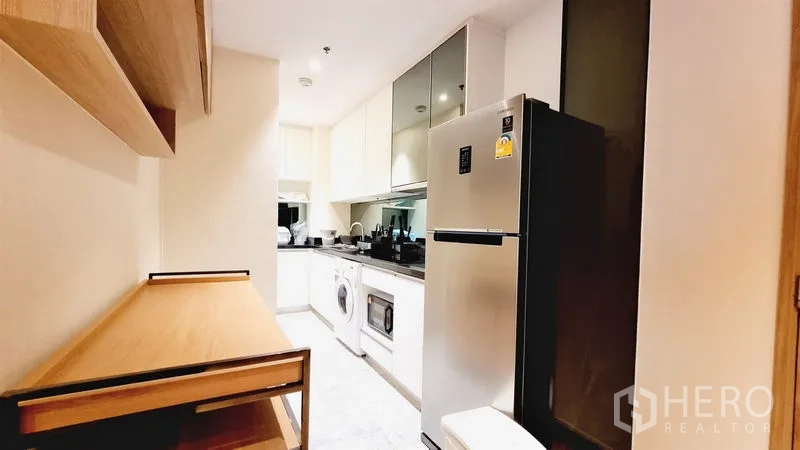 Condominium for sale in Watthana, Bangkok - Bright galley kitchen with glossy white cabinets, black stone countertop, stainless fridge, washer and microwave in a Phra Khanong condo.