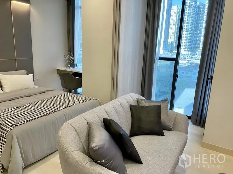 Condominium for rent in Khlong Toei, Bangkok - Bedroom and lounge area with sofa, work desk and floor-to-ceiling windows showing city skyline.